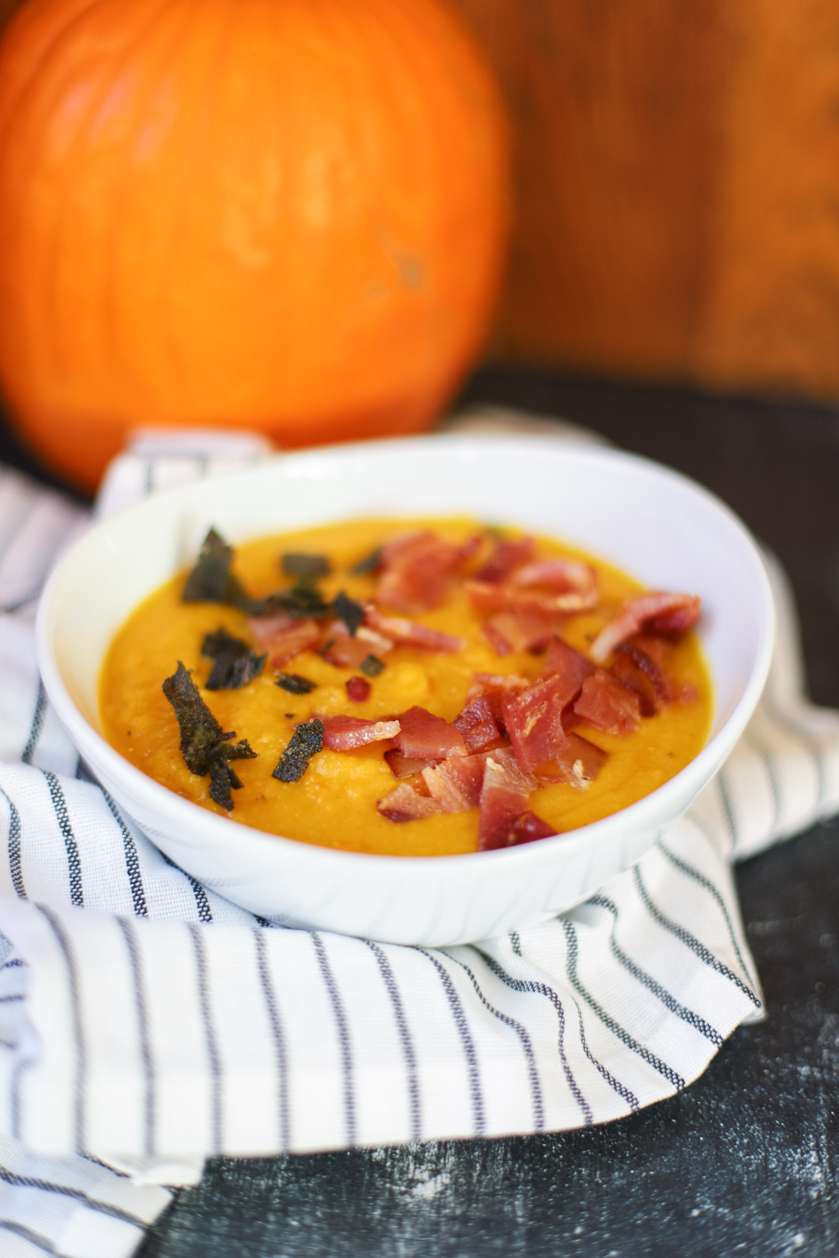 Creamy carrot pumpkin soup topped with crispy bacon and fried sage, with a pumpkin in the background.