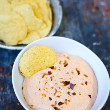 A bowl of creamy cottage cheese queso dip topped with crushed red pepper flakes, served with tortilla chips in the background