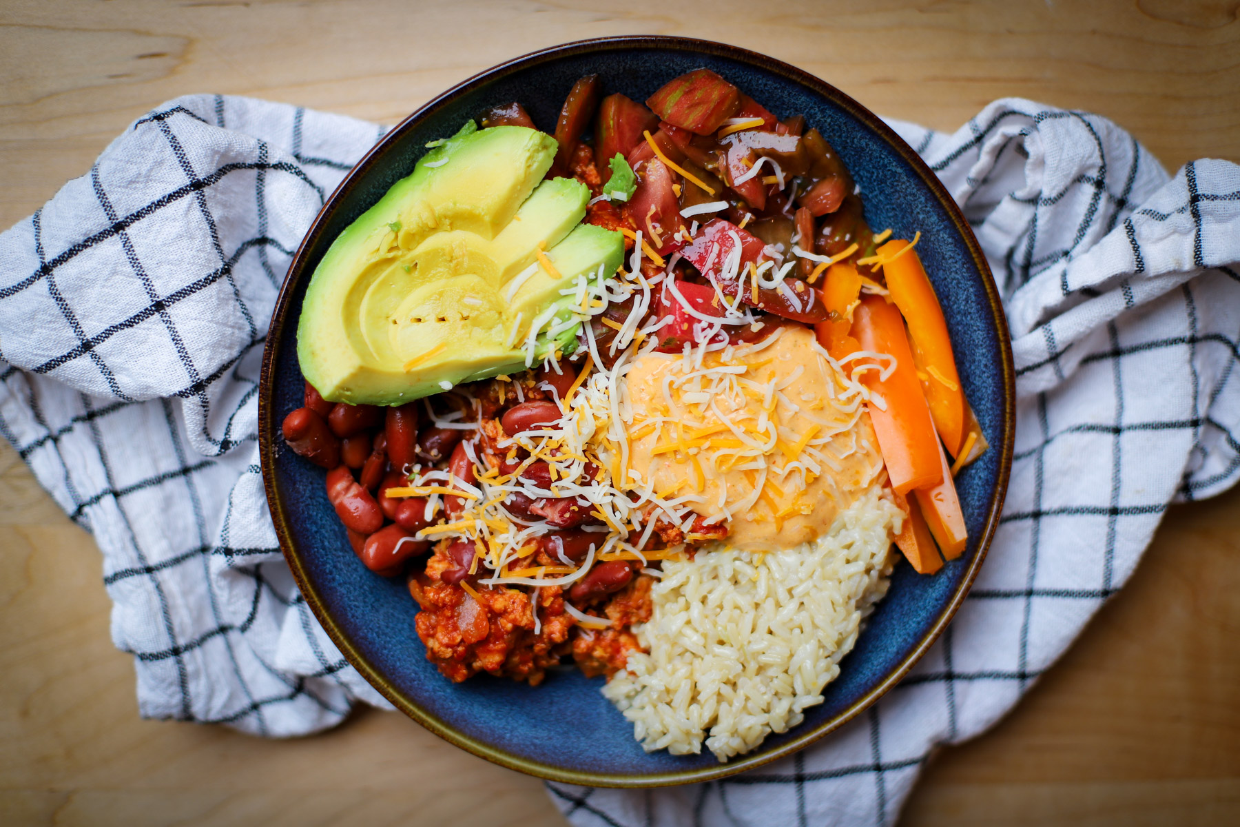 A colorful grain bowl topped with cottage cheese queso, seasoned ground meat, rice, kidney beans, shredded cheese, avocado slices, and fresh vegetables.