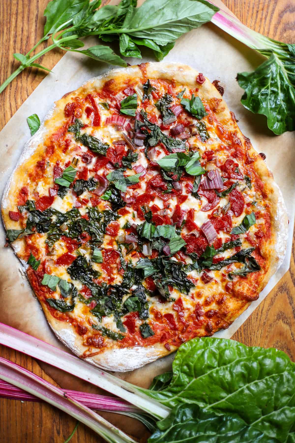 A Swiss chard and pepperoni pizza surrounded by Swiss chard and basil on a cutting board