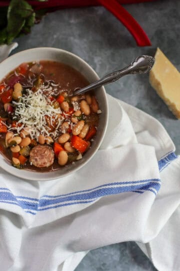 Close-up of a bowl of slow cooker andouille sausage stew featuring Swiss chard, white beans, carrots, and shredded Parmesan cheese on a blue-striped white cloth with a wedge of Parmesan on a gray marble surface.