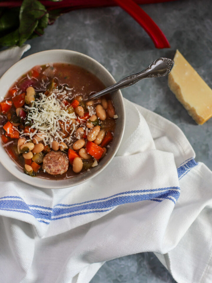 Close-up of a bowl of slow cooker andouille sausage stew featuring Swiss chard, white beans, carrots, and shredded Parmesan cheese on a blue-striped white cloth with a wedge of Parmesan on a gray marble surface.