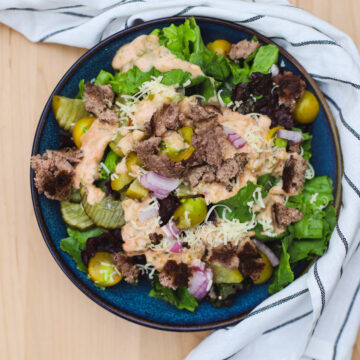 Big Mac salad in a blue bowl with fresh lettuce, pickles, ground beef, shredded cheese, onions, and special sauce drizzle, placed on a wooden surface with a striped napkin.