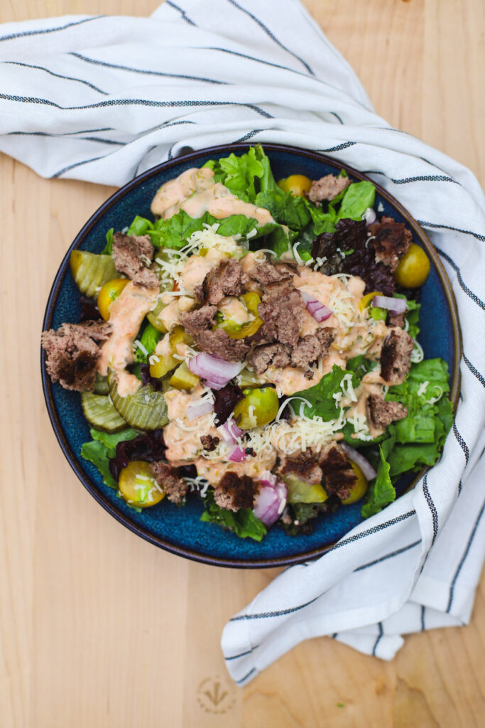 Big Mac salad in a blue bowl with fresh lettuce, pickles, ground beef, shredded cheese, onions, and special sauce drizzle, placed on a wooden surface with a striped napkin.