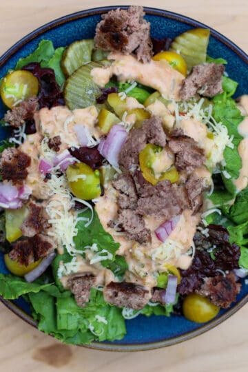Close-up of Big Mac salad in a blue bowl with ground beef, lettuce, pickles, shredded cheese, diced red onions, and special sauce on a wooden background.
