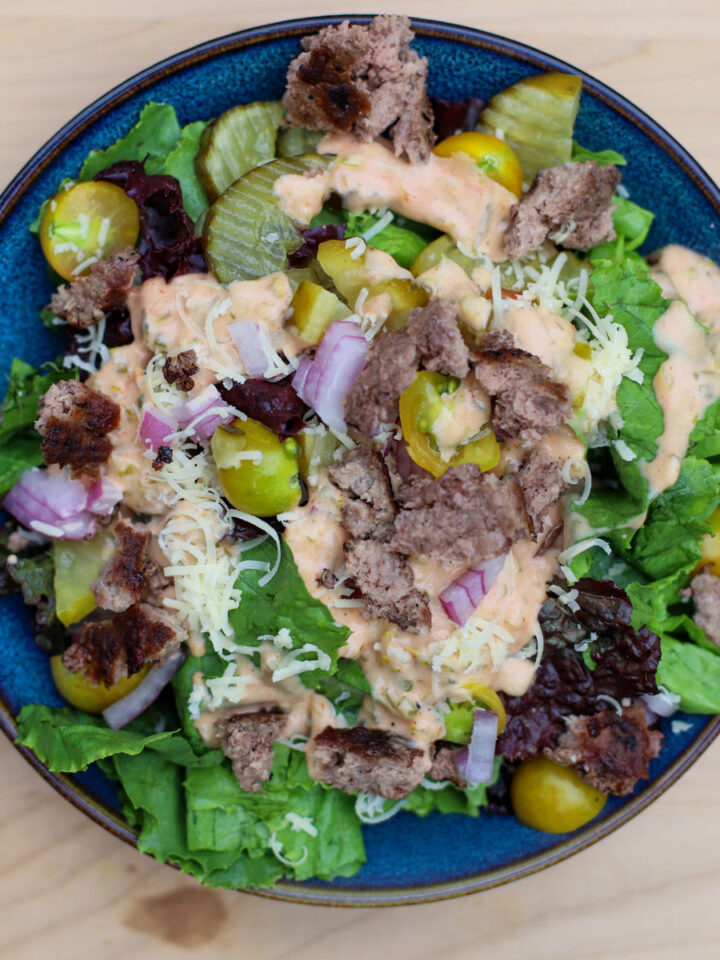 Close-up of Big Mac salad in a blue bowl with ground beef, lettuce, pickles, shredded cheese, diced red onions, and special sauce on a wooden background.