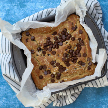 Chickpea blondies with chocolate chips in a parchment-lined baking pan on a striped towel, set against a textured blue background.