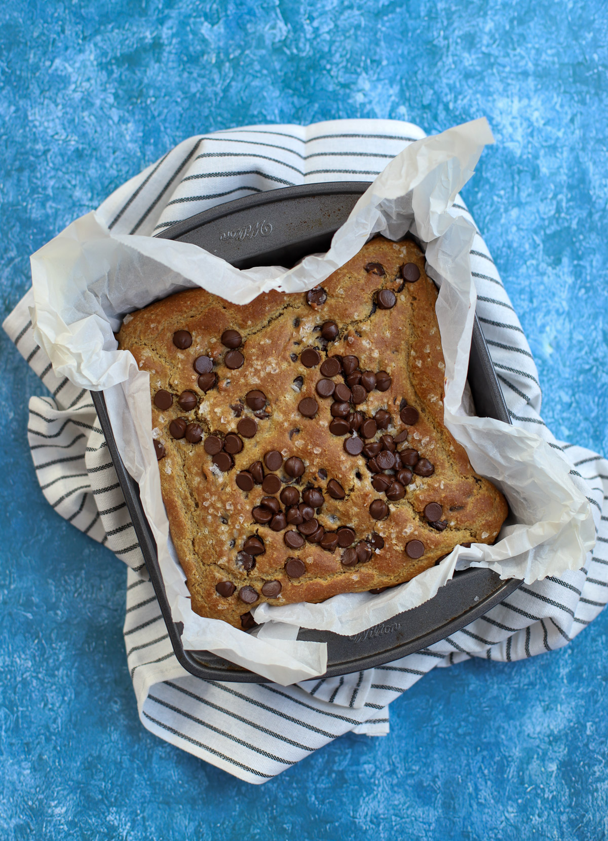 Freshly baked chickpea blondies topped with chocolate chips in a parchment-lined baking pan, placed on a striped towel against a blue background.