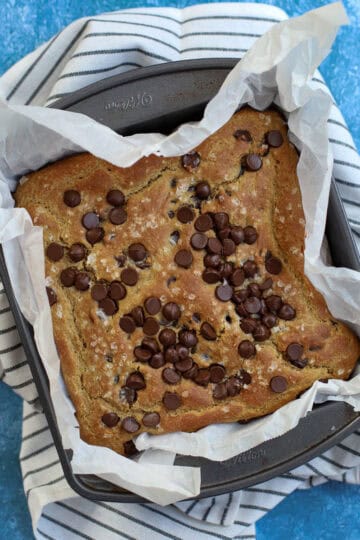 Close-up of chickpea blondies topped with chocolate chips, baked in a parchment-lined pan on a striped towel with a blue textured background.