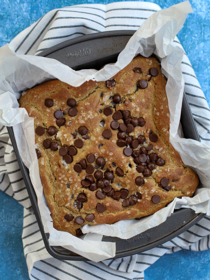 Close-up of chickpea blondies topped with chocolate chips, baked in a parchment-lined pan on a striped towel with a blue textured background.