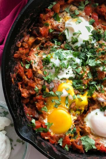 Close-up of shakshuka in a cast iron skillet, featuring perfectly poached eggs nestled in a spiced tomato and vegetable sauce, topped with fresh cilantro, and served alongside a bowl of yogurt.