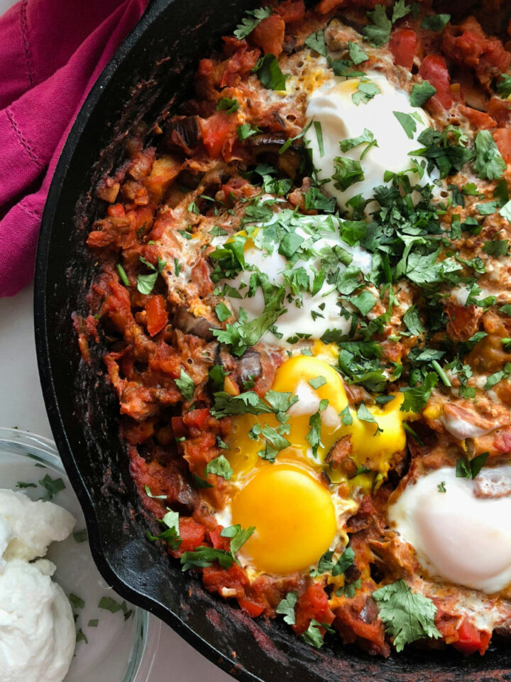 Close-up of shakshuka in a cast iron skillet, featuring perfectly poached eggs nestled in a spiced tomato and vegetable sauce, topped with fresh cilantro, and served alongside a bowl of yogurt.