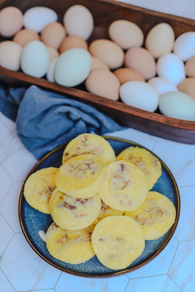 Plate of homemade cottage cheese egg bites with bacon, next to a rustic wooden bowl filled with farm-fresh eggs on a tiled countertop.