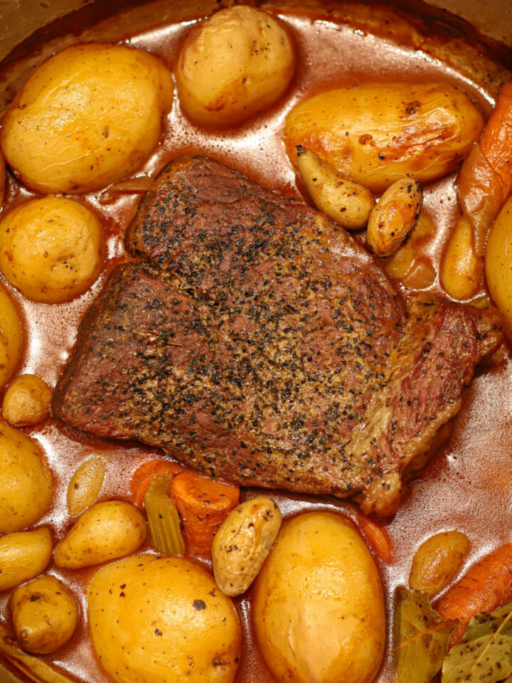 Close-up of a Dutch oven pot roast with golden potatoes, carrots, celery, and bay leaves simmering in a flavorful broth.