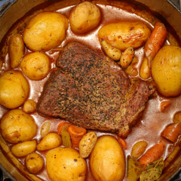 Tender Dutch oven pot roast surrounded by golden potatoes, carrots, celery, and bay leaves in a rich broth, cooked on the stovetop.