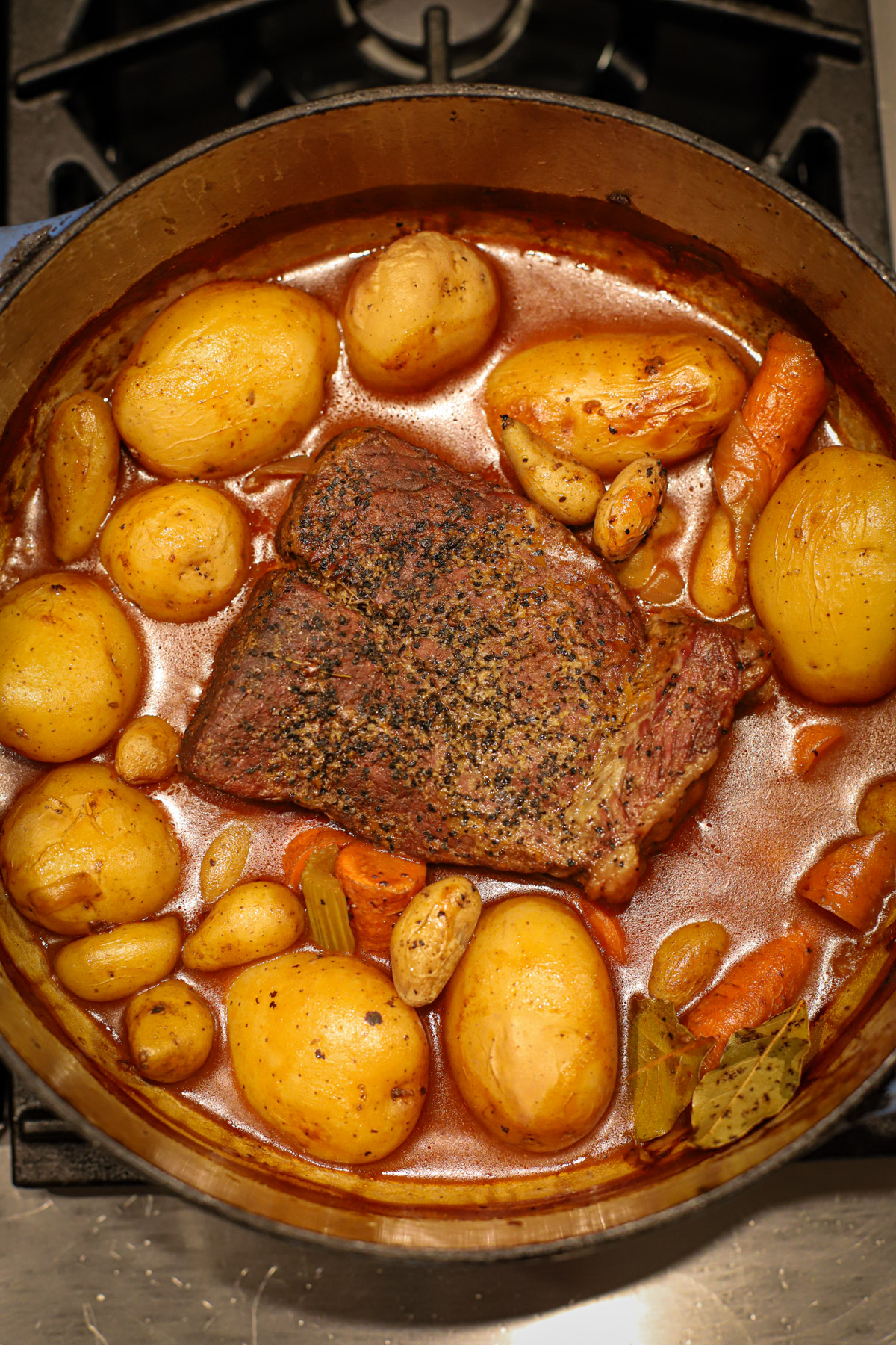 Tender Dutch oven pot roast surrounded by golden potatoes, carrots, celery, and bay leaves in a rich broth, cooked on the stovetop.