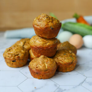 Golden zucchini carrot muffins stacked on a hexagon-tiled surface with fresh ingredients like zucchini, eggs, and carrots in the background.