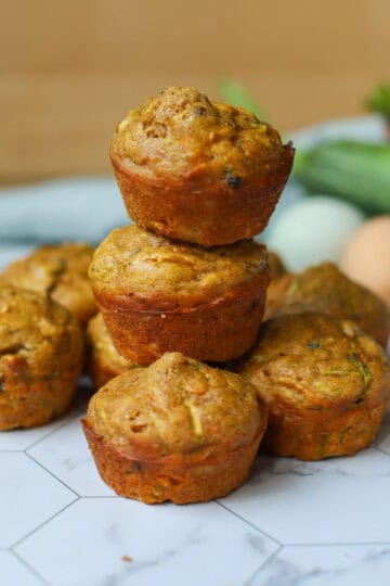 Golden zucchini carrot muffins stacked on a hexagon-tiled surface with fresh ingredients like zucchini, eggs, and carrots in the background.