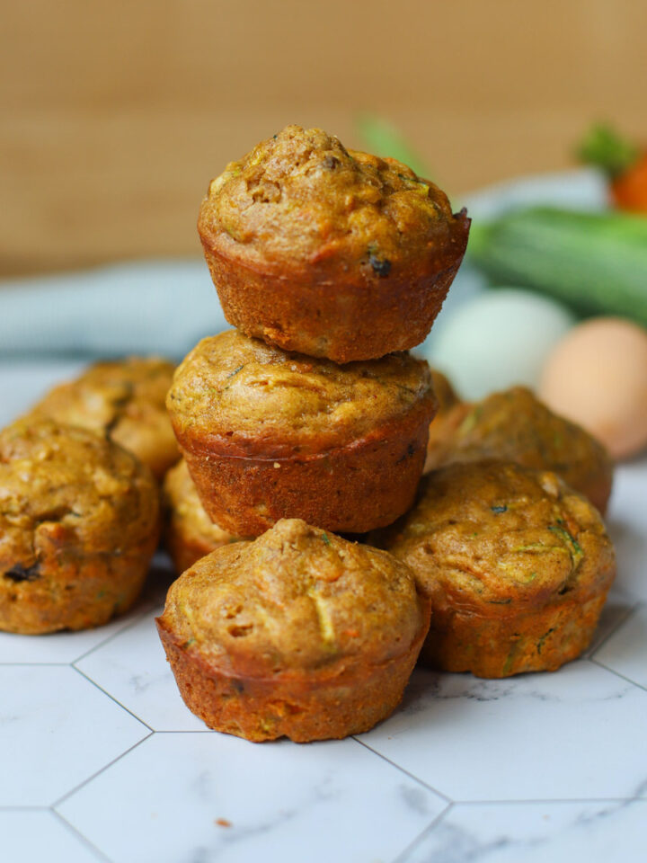 Golden zucchini carrot muffins stacked on a hexagon-tiled surface with fresh ingredients like zucchini, eggs, and carrots in the background.