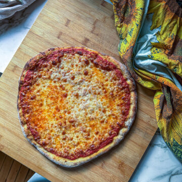 Homemade cheese pizza with a golden, bubbly crust, topped with a simple tomato sauce and melted cheese, on a wooden board with a sunflower-patterned towel nearby.