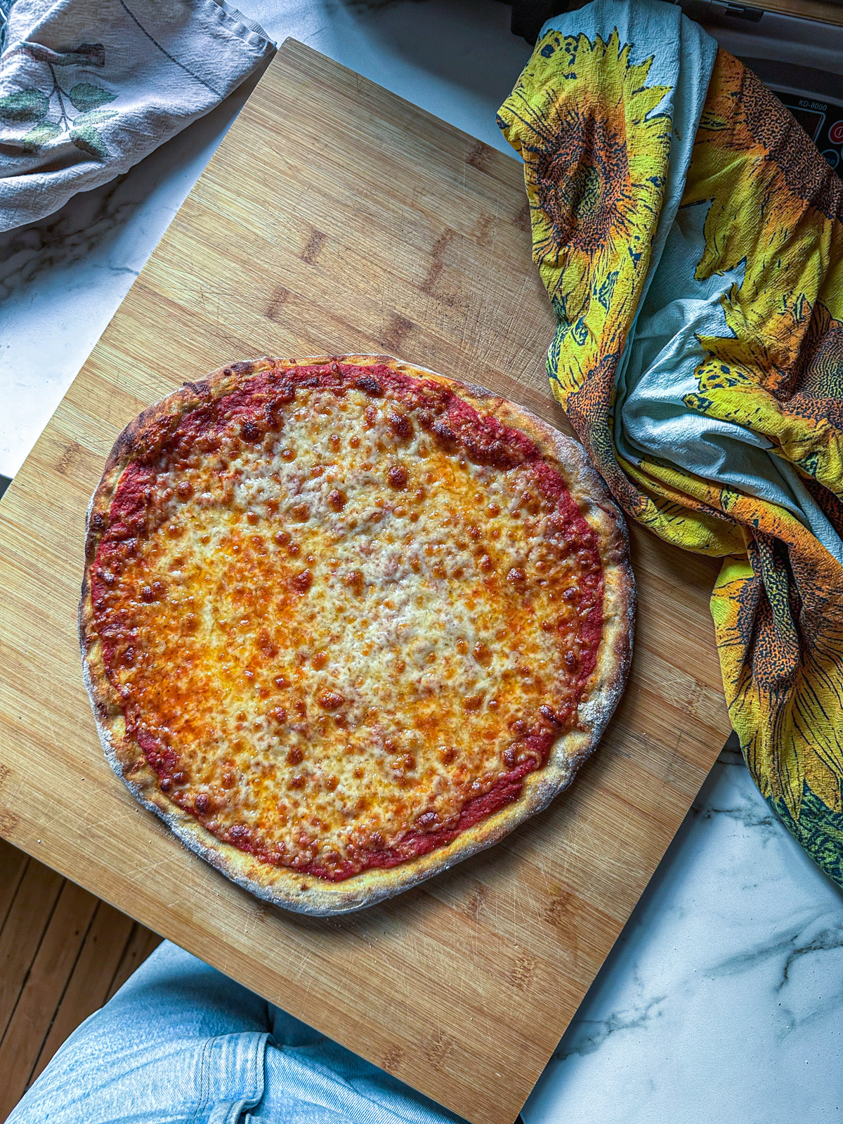 Homemade cheese pizza with a golden, bubbly crust, topped with a simple tomato sauce and melted cheese, on a wooden board with a sunflower-patterned towel nearby.