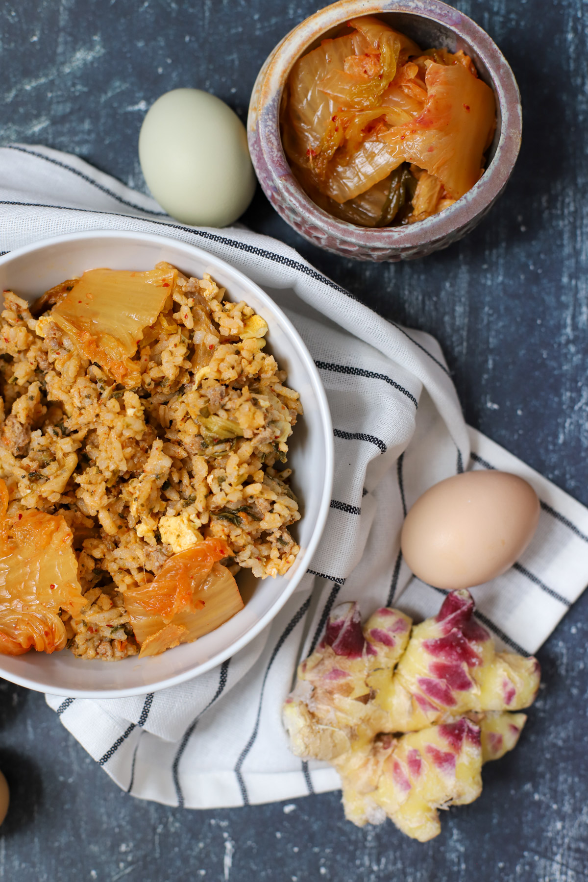 Close-up of a bowl of kimchi rice with pork and cabbage, topped with kimchi leaves, surrounded by fresh eggs, ginger, and a jar of kimchi on a striped napkin.