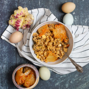 Styled overhead shot of a bowl of kimchi rice with pork and cabbage, garnished with kimchi leaves, surrounded by farm-fresh eggs, purple ginger, and a jar of kimchi on a striped cloth.