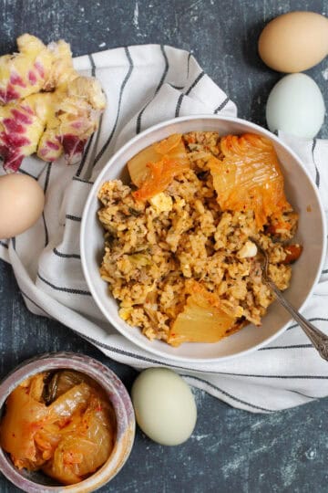 Styled overhead shot of a bowl of kimchi rice with pork and cabbage, garnished with kimchi leaves, surrounded by farm-fresh eggs, purple ginger, and a jar of kimchi on a striped cloth.