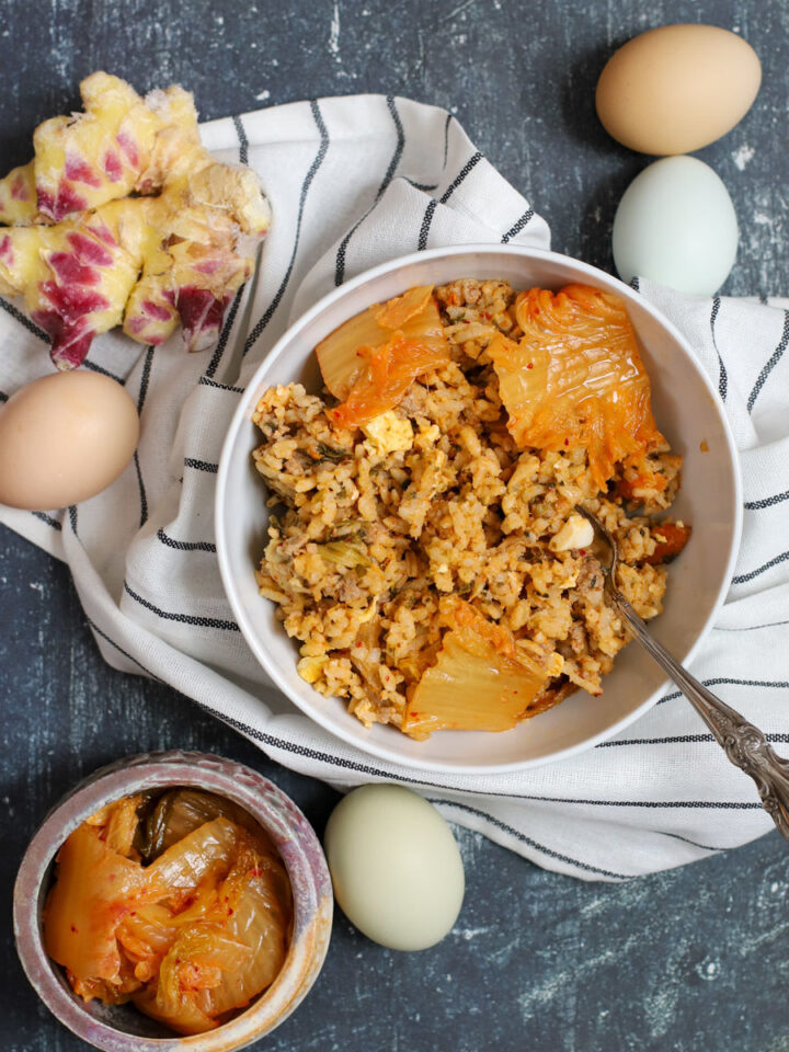 Styled overhead shot of a bowl of kimchi rice with pork and cabbage, garnished with kimchi leaves, surrounded by farm-fresh eggs, purple ginger, and a jar of kimchi on a striped cloth.