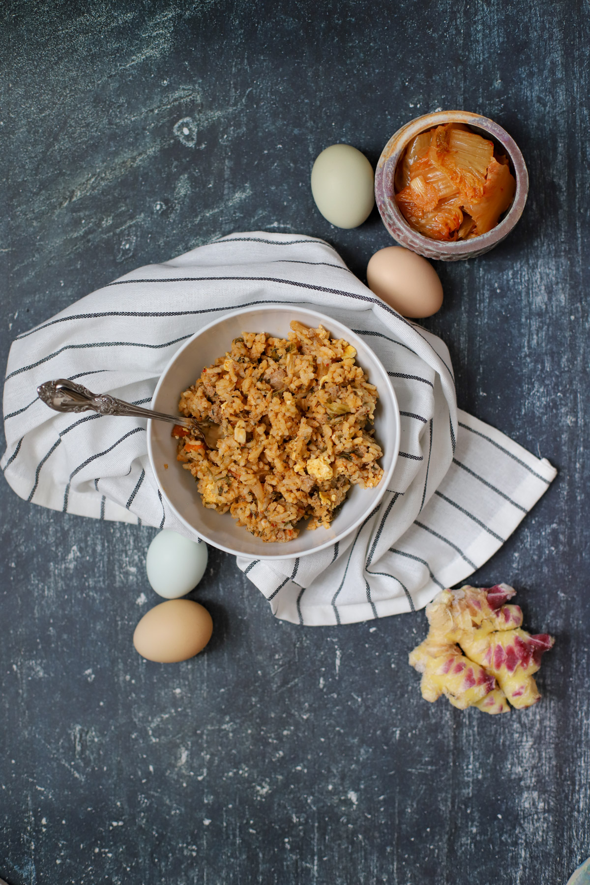 Bowl of spicy kimchi rice with pork, cabbage, and scrambled egg on a striped napkin, surrounded by fresh eggs, kimchi, and ginger.