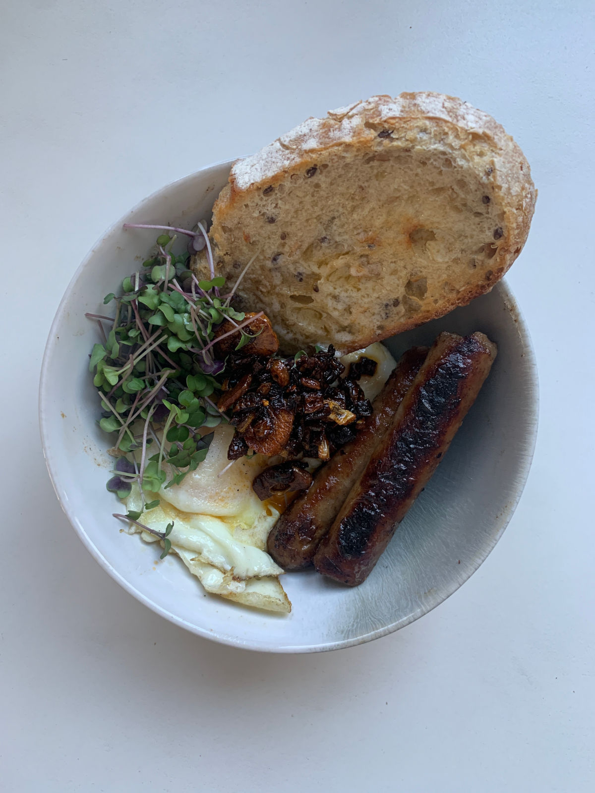 Breakfast bowl featuring fresh microgreens, sausages, fried egg, roasted vegetables, and a slice of artisan bread.