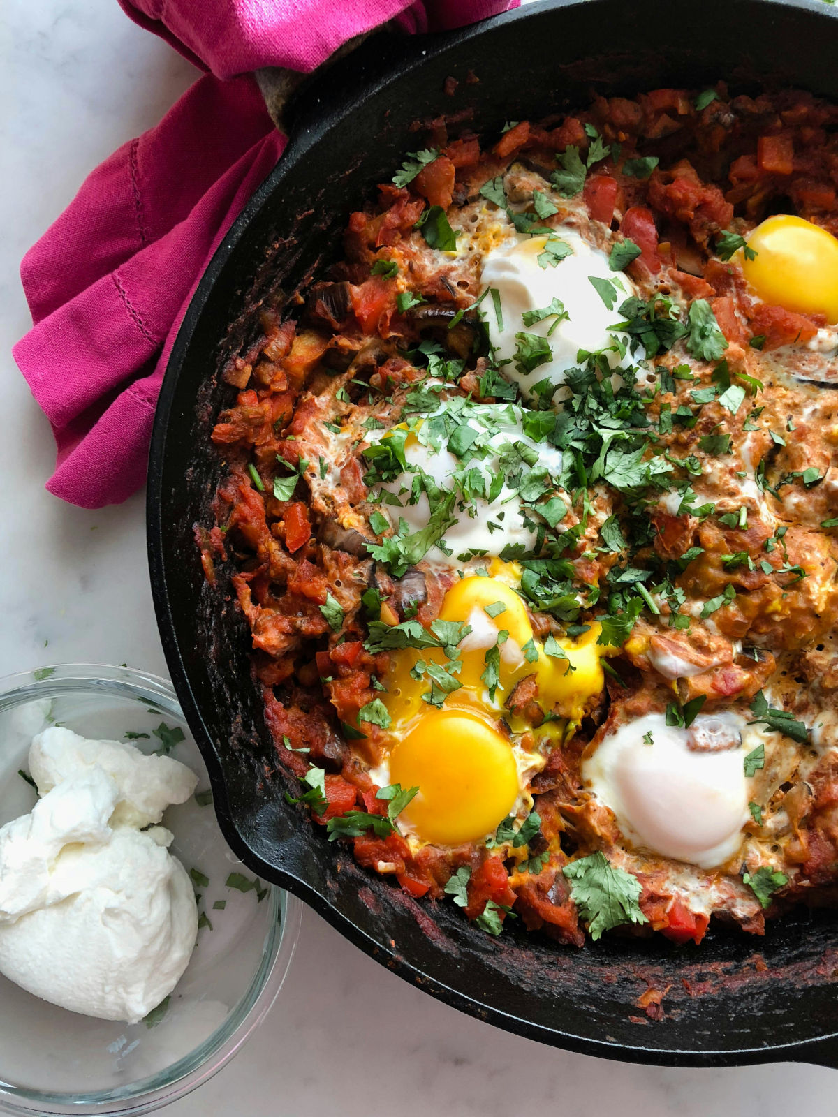 Close-up of a cast iron skillet filled with shakshuka, featuring poached eggs in a rich tomato and vegetable sauce, garnished with fresh cilantro, and served with a side of creamy yogurt on a marble countertop.