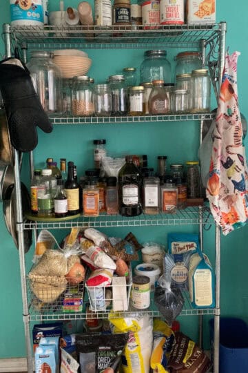 Close-up view of a well-organized pantry with shelves stocked with essential ingredients like grains, canned goods, oils, spices, and baking supplies against a teal wall.