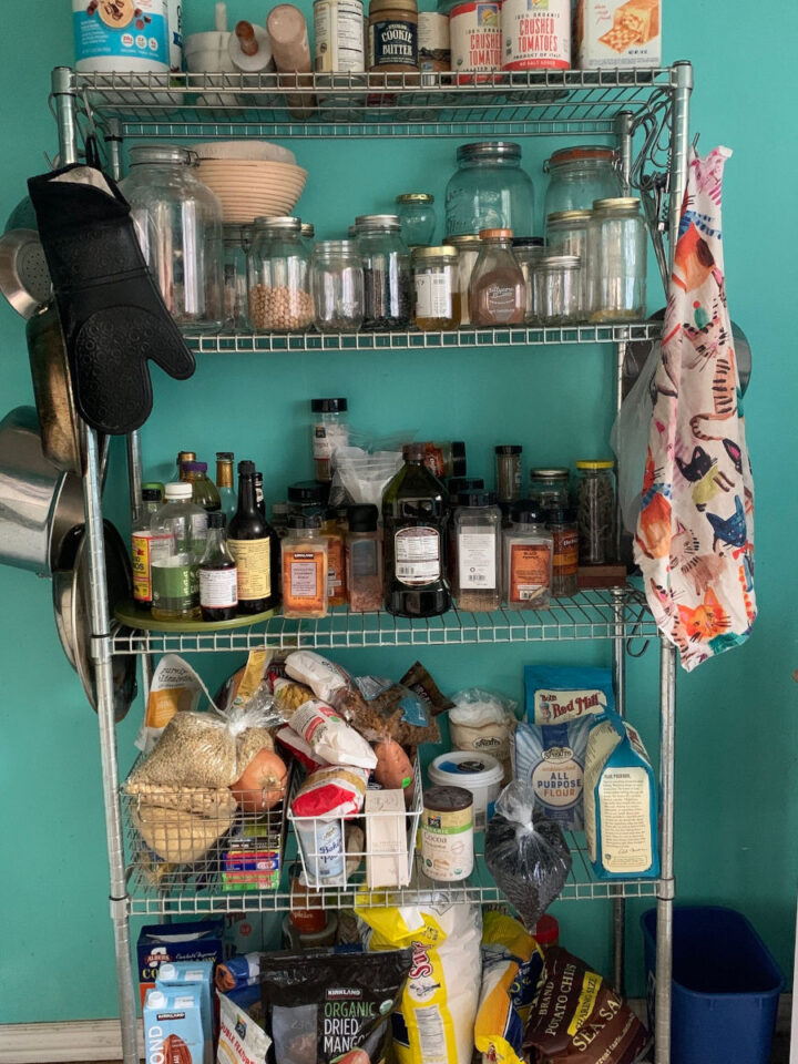 Close-up view of a well-organized pantry with shelves stocked with essential ingredients like grains, canned goods, oils, spices, and baking supplies against a teal wall.