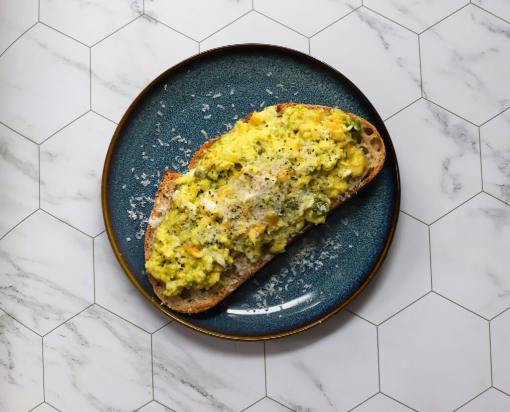 Overhead shot of pesto scrambled eggs on toasted sourdough bread, garnished with grated Parmesan and cracked black pepper, served on a dark blue plate on a marble hexagon tile background.