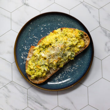 Overhead shot of pesto scrambled eggs on toasted sourdough bread, garnished with grated Parmesan and cracked black pepper, served on a dark blue plate on a marble hexagon tile background.