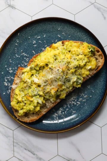 Close-up of pesto scrambled eggs topped with Parmesan and black pepper on a slice of sourdough toast, displayed on a dark blue plate with a marble hexagon tile background.