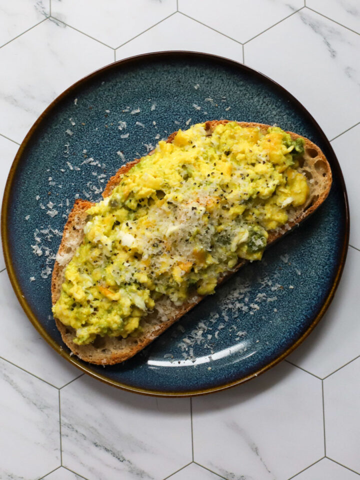 Close-up of pesto scrambled eggs topped with Parmesan and black pepper on a slice of sourdough toast, displayed on a dark blue plate with a marble hexagon tile background.