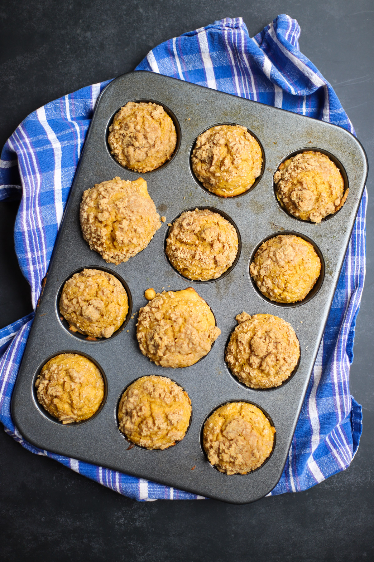 Freshly baked pumpkin streusel muffins in a muffin tin, placed on a blue plaid towel, showcasing golden brown tops with crumbly streusel.