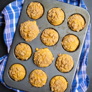 Batch of freshly baked pumpkin streusel muffins in a muffin tin, with crumbly golden-brown topping on each muffin and a blue plaid towel underneath.