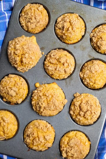 Batch of freshly baked pumpkin streusel muffins in a muffin tin, with crumbly golden-brown topping on each muffin and a blue plaid towel underneath.