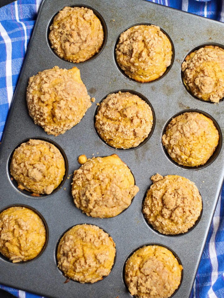 Batch of freshly baked pumpkin streusel muffins in a muffin tin, with crumbly golden-brown topping on each muffin and a blue plaid towel underneath.