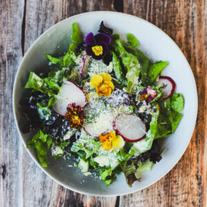 Close-up of fresh summer salad with mixed greens, radishes, grated parmesan, and colorful edible flowers on a rustic wood table.