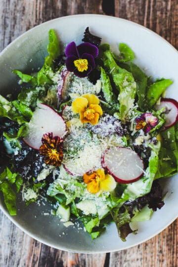 Close-up of fresh summer salad with mixed greens, radishes, grated parmesan, and colorful edible flowers on a rustic wood table.