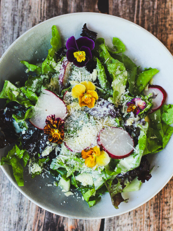 Close-up of fresh summer salad with mixed greens, radishes, grated parmesan, and colorful edible flowers on a rustic wood table.