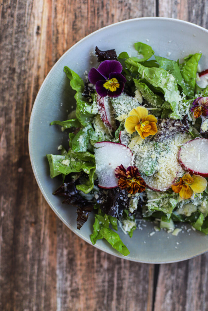 Fresh summer salad with mixed greens, radish slices, parmesan, and edible flowers on a rustic wood table.