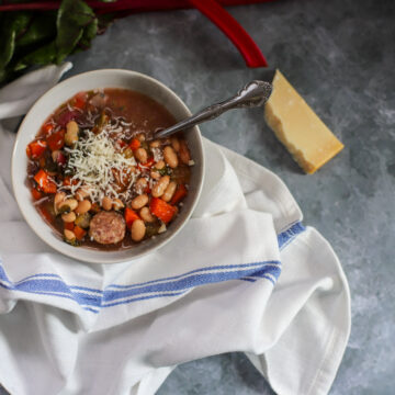 Bowl of slow cooker andouille sausage stew with Swiss chard, white beans, and carrots, topped with shredded Parmesan cheese, styled with a blue-striped white cloth on a gray marble surface.