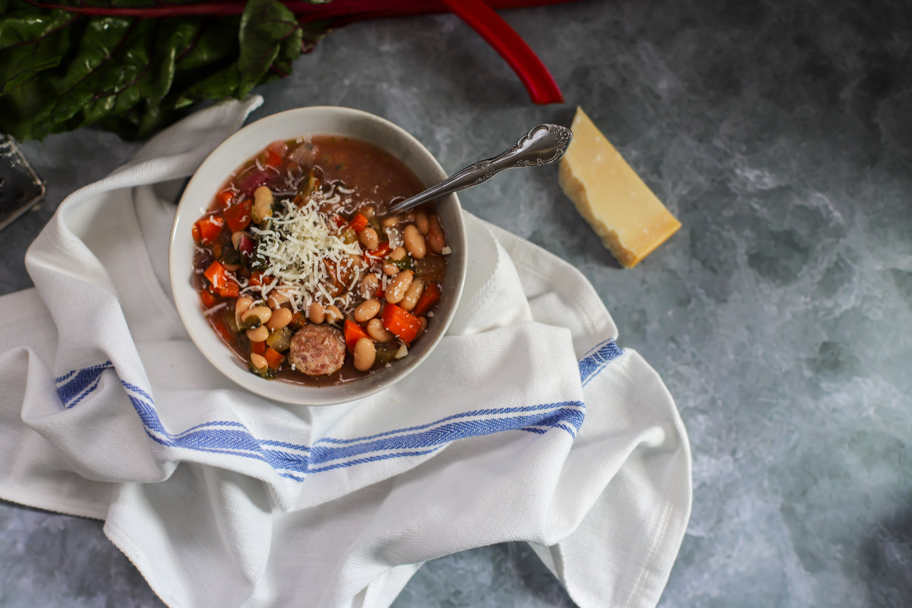 Bowl of slow cooker andouille sausage stew with Swiss chard, white beans, and carrots, topped with shredded Parmesan cheese, styled with a blue-striped white cloth on a gray marble surface.