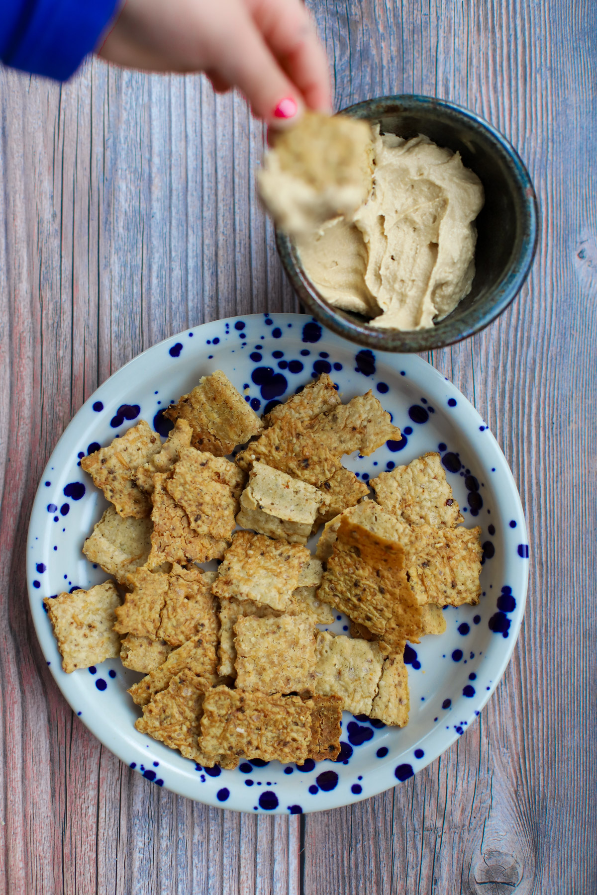 Homemade sourdough discard crackers on a blue and white speckled plate, with a child dipping one into a bowl of creamy hummus.