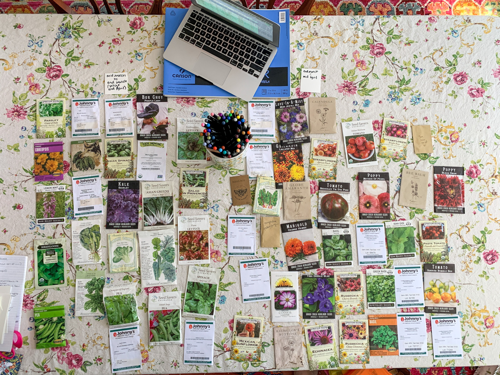 A large collection of seed packets displayed on a floral tablecloth, including vegetables, herbs, and flowers from trusted seed companies like Johnny's Selected Seeds, Seed Savers Exchange, and Baker Creek Heirloom Seeds.